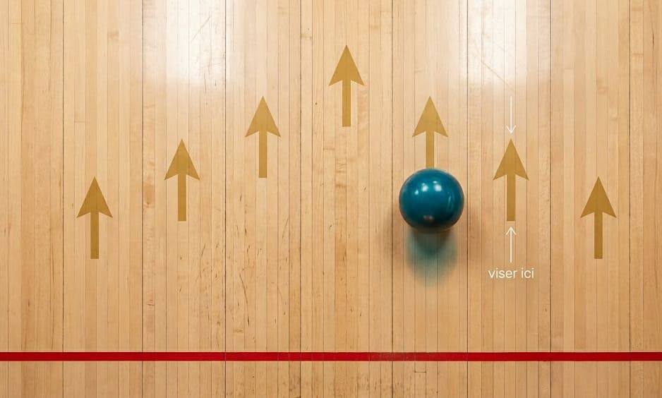 Vue zénithale éditoriale d'une piste de bowling, sept flèches dorées au sol, boule cyan posée sur la deuxième flèche depuis la droite