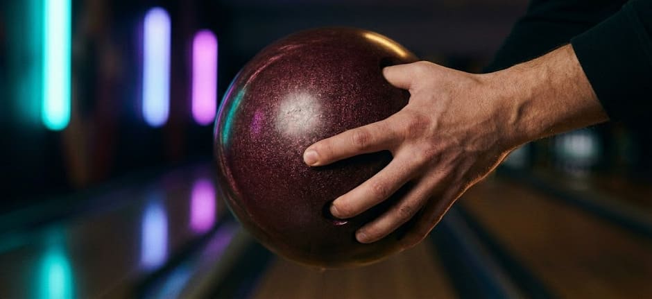 Macro éditoriale : prise conventionnelle d'une boule de bowling, focus sur les doigts insérés dans les trous, fond néon en bokeh