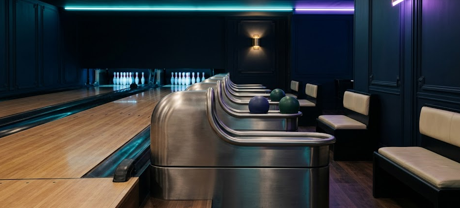 Vue rasante du couloir entre deux pistes de bowling parisien : retours de boules en métal brossé et néons cosmic